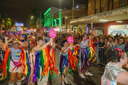 Group of smiling and dancing people with rainbow coloured ribbon costumes and signs wave at crowds of people during Sydney Mardi Gras