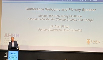 Photo of a man speaking at a lectern, with a big screen behind him that says "Conference Welcome and Plenary Speaker"