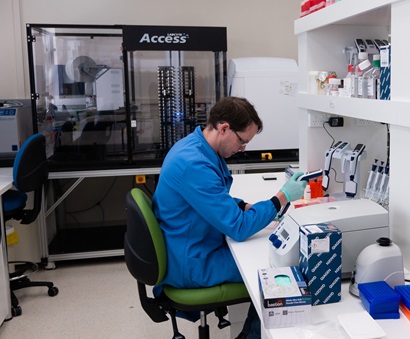 A man in a blue coat sitting at a laboratory bench working on samples with the acoustic liquid handler in the background.