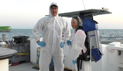 Two people sanding on a boat deck, wearing white overalls, one with a large blue backpack.