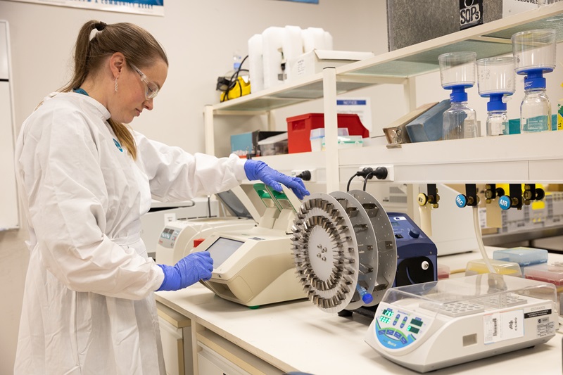 A scientist standing at a laboratory bench in front of a PCR machine, loading a sample.