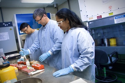 Three people in blue labcoats standing at a bench in a laboratory.