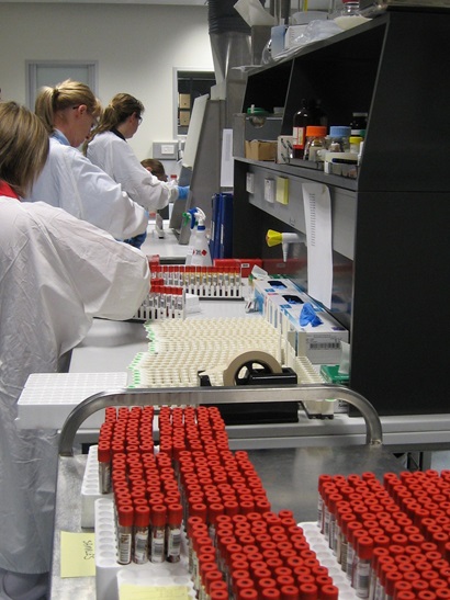 Trays of rows of red coloured sample test tubes on a bench with three people in white lab coats in the background.