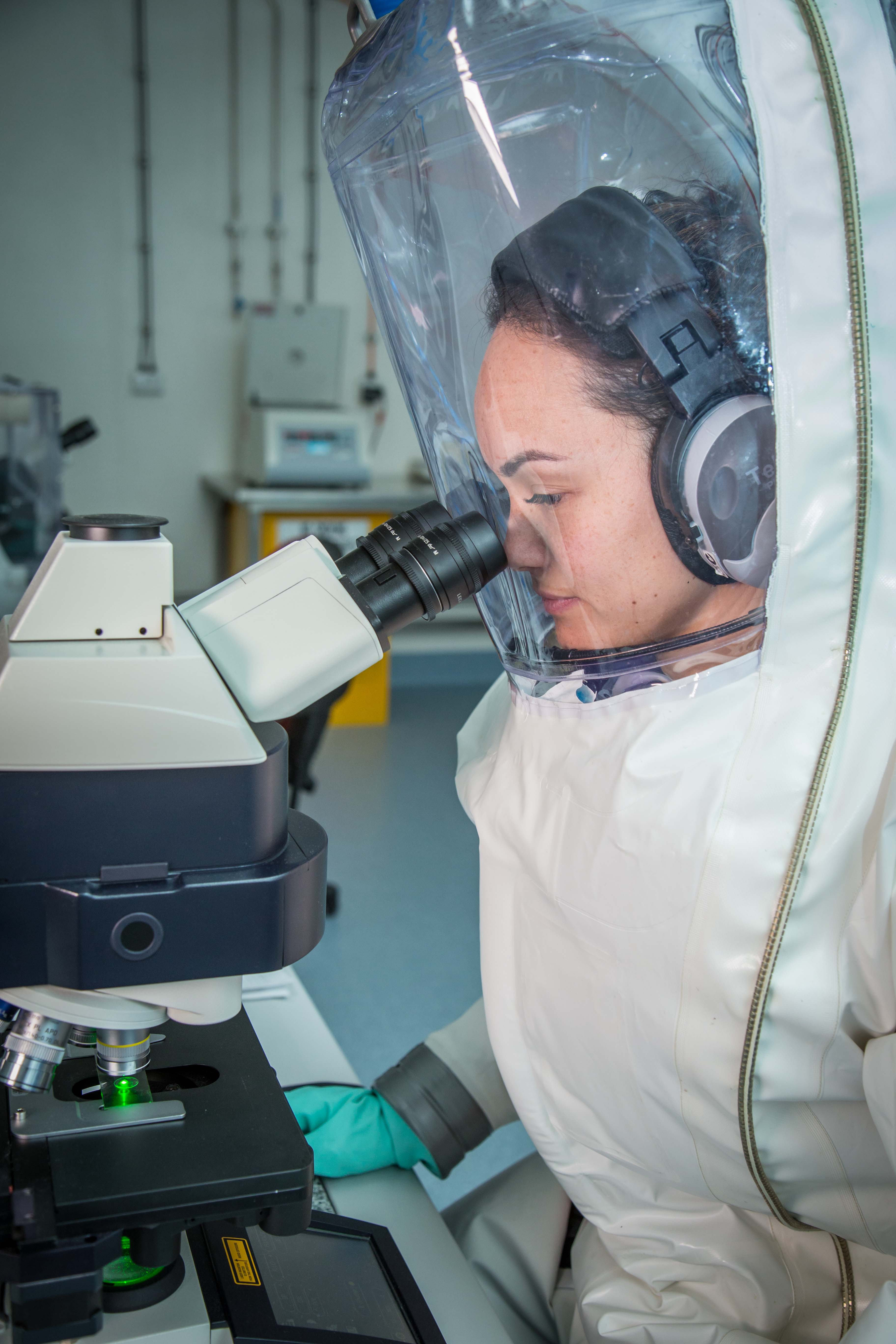 A woman in a fully enclosed suit connected to a separate air supply sitting at a bench looking into a microscope.