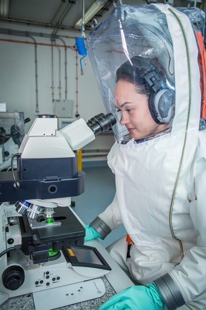 A women in a fully enclosed protective suit, sitting at a microscope at a bench in a laboratory.