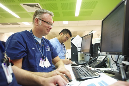 A doctor sitting in front of a computer screen with other medical staff in the background of a hospital room