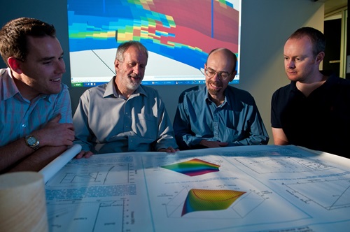 Four male researchers, Martin Leahy, Lincoln Paterson, Jonathan Ennis-King and Chris Green, seated around table covered with research diagrams with projection of geological model in background.