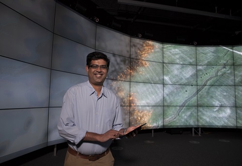 Dr Mahesh Prakash holding a computer tablet in front of a bank of screens showing Sparks software in use.