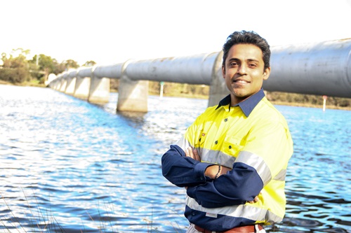 Man in high vis clothes standing next to a body of water with large pipeline running through the background. 