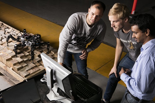 Three scientists working at a computer testing a hexapod robot on a side table.