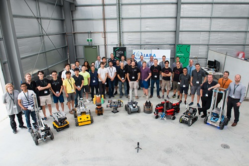 In a warehouse, a large group of people stand in a semi-circle with several small robots positioned in front of them.