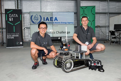 Two men kneel behind a caterpillar tracked robotic device. 