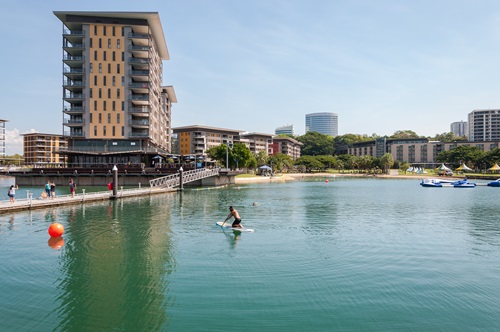Waterfront view with buildings in the background and people swimming and paddleboarding in the water. 