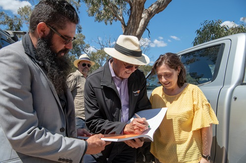 Trent Nelson and Graham Atkinson standing with the Hon Lily D'Ambrosio MP next to a vehicle and holding a copy of the Joint Management Plan.