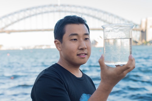Person holding a beaker of water taken from Sydney Harbour, with the Sydney Harbour Bridge in the backgorund. 