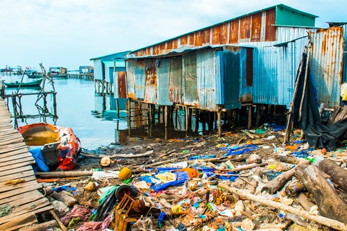 Section of shoreline covered with rubbish with wooden dock on one side and hut on the other. 