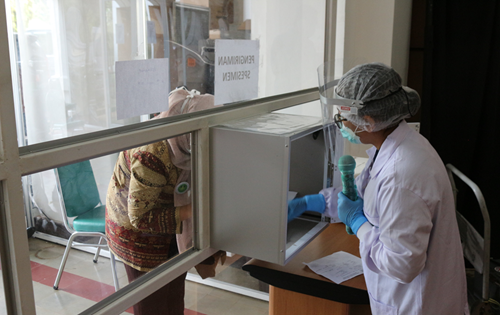 Person working in protective coverings behind a glass partition wall, collecting specimen samples through a small opening.  