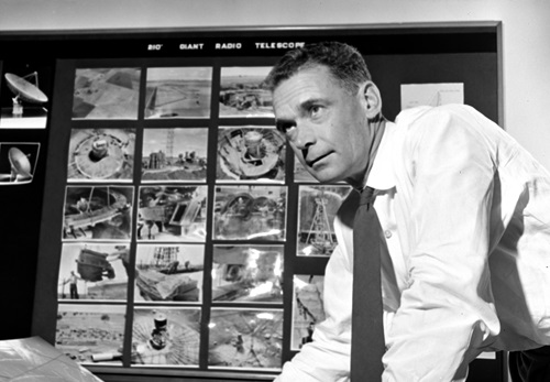 Dr E.G. 'Taffy' Bowen leaning over a desk with photos of the Parkes telescope in various stages of construction pinned to a board in the background. 