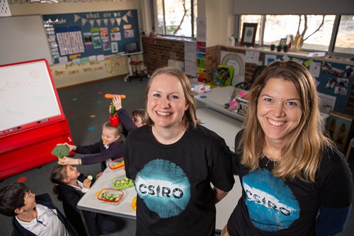 Two scientists stand in front of children eating vegetables