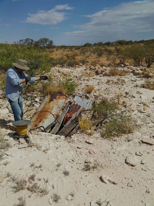 Person sampling water from the opening of an underground well covered in corrugated iron in bushland. 