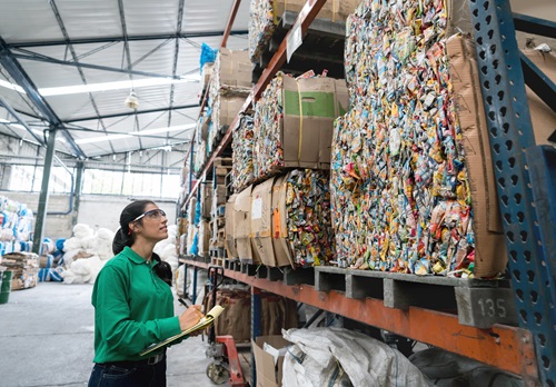 Woman holding a clipboard looking up at large palettes of packed rubbish