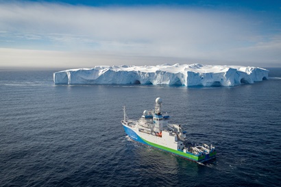 Drone photo of the RV Investigator with a large iceberg in the background