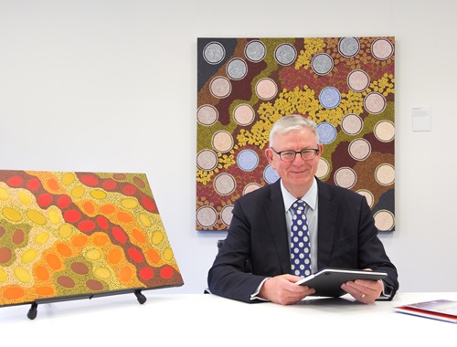 A man in a suit, holding an electronic notepad, sits next to a decorative painting. Another decorative painting is behind him on the wall.