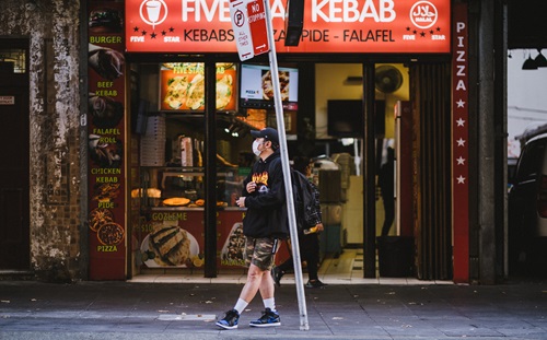 Person wearing facemask, leaning against a street sign in front of a kebab shop. 
