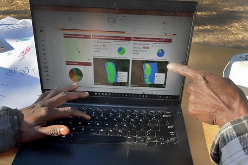 Hand of one person working at a laptop, while the hand of another points at something on the screen. 