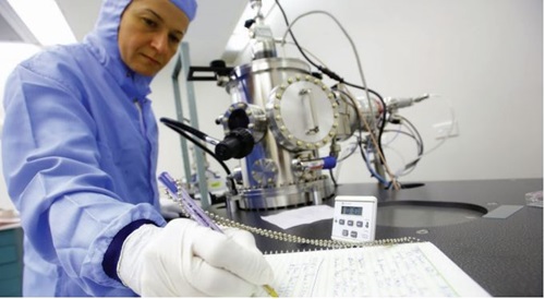 Researcher in purple laboratory coat and white gloves standing and writing notes near industrial science machinery.