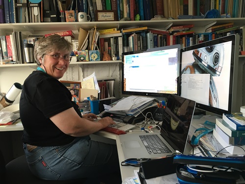 Woman with short silver seated at desk in front of computer screens