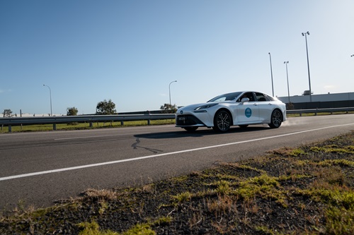 Image of a white Toyota hybrid car driving on a bitumen road bordered grass and a silver guard rail in the background.