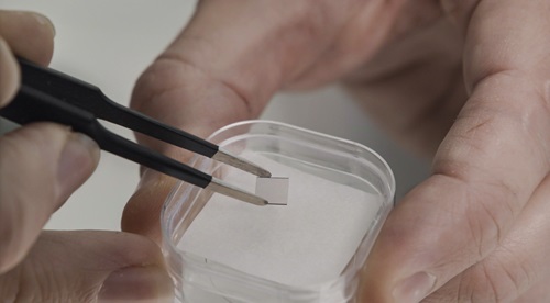 Tweezers pick up a small silver square from a small padded box held between a thumb and finger