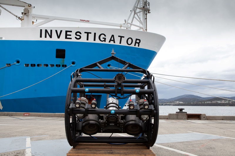 The deep-tow camera rig waits on the wharf, in front of a big blue and white ship called the Investigator. On the rig, there are multiple cameras and lights secured and wired.