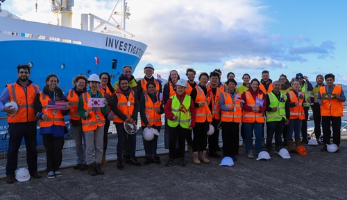 A group of people in orange and yellow hi-vis vests stand in front of a ship.
