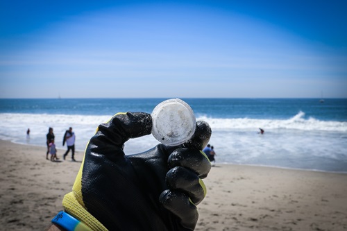 Hand wearing a black glove holds up a bottle lid found as rubbish with the beach in the background