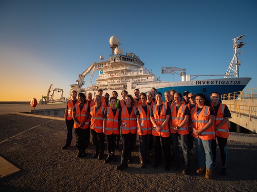 A group of people in hi vis vests standing in front of a ship at sunset.