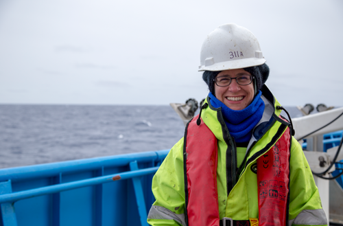 A person in cold weather gear and a white hard hat standing on the deck of a ship with the ocean in the background.