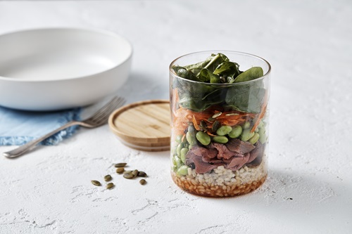 A round glass jar sits open on a white bench. In the jar is a layered salad. Starting with the base layer there is brown rise with a red sauce, the next layer is beef strips and green edamame beans. The top layer is grated carrot and baby spinach leaves. In the background there is an empty white bowl and a fork with the jar’s wooden lid and a few pepita seeds sprinkled next to the jar.    