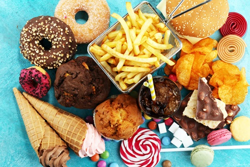 A photograph of an assortment of different junk foods taken from above including donuts, ice cream cones, muffins, fried chips, a glass with cola soft drink, chocolate, macaroons, smarties, a lollypop, colour liquorice and a hamburger.    