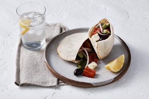  A white and brown ceramic plate sits in the foreground. On it is a pita pocket cut in half which contains cut cucumber, black olives, red onions, red and green capsium and feta cheese. On the slide of the plate is a lemon wedge. In the background of the photos is a glass of water with ice cubes and cut lemon pieces. The glass sits on a sand coloured linen napkin.     