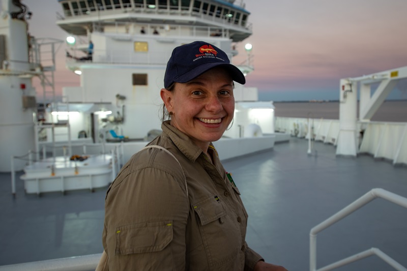 Photo of a woman (Dr Kate Grarock) smiling at the camera. She is standing on the deck of a research vessel (RV Investigator).
