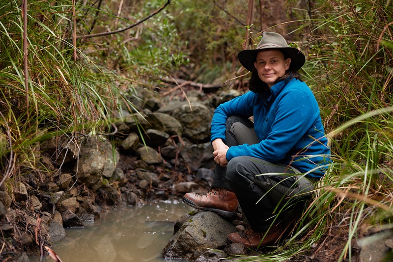 A promotional photo of a woman (Dr Kate Grarock) on sitting near a small creek in Tasmania as part of the television show Alone Australia.