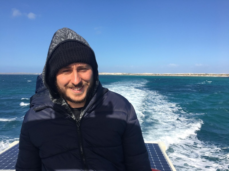 Photo of a young man in a beanie and warm jacket on a boat in the sun. Solar panels and the ocean can be seen behind him.