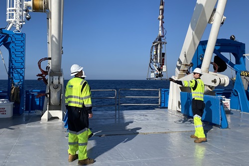 Three people in hi-vis deploy a piece of scientific equipment from the deck of a ship.