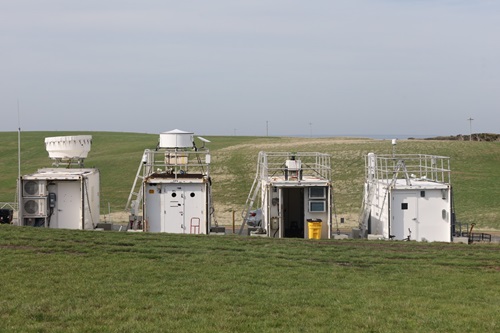 Four white containers in a row sitting on a grassy hill. The sky is overcast. There is instrumentation and equipment attached to each container.