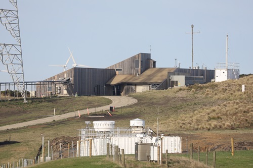 The Kennaook / Cape Grim Baseline Air Pollution station, from a distance. The white ARM mobile laboratory is sitting just below the station. There is a mobile phone tower just to the left of the station, and wind turbines in the background.