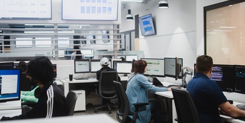 A modern office environment with multiple people working at computer workstations. Each person is focused on their screens, which display various data, graphs, and code. The room is well-lit with overhead lights and has several large monitors mounted on the walls showing charts and graphs. The workspace is organised with ergonomic chairs and desks equipped with multiple monitors for each individual.