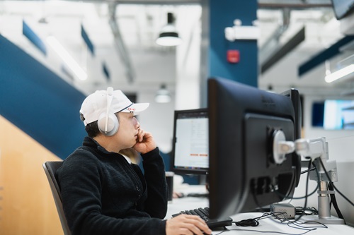 A person wearing a white cap and large headphones is sitting at a desk in an office environment. The person is facing a computer monitor, with one hand resting on their chin. The background shows an open office space with modern lighting and blue walls. The computer screen displays some text, but it is not clearly readable in the image. 