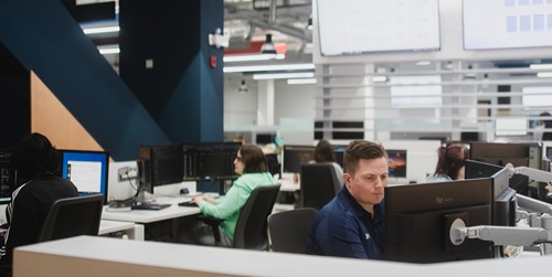 The image shows a modern office environment with several people working at their desks. Each person is focused on their computer screens, which display various software and data. The office has a clean, organised layout with multiple monitors and ergonomic chairs. The background includes large screens mounted on the wall displaying charts and graphs, indicating a data-driven work setting. 
